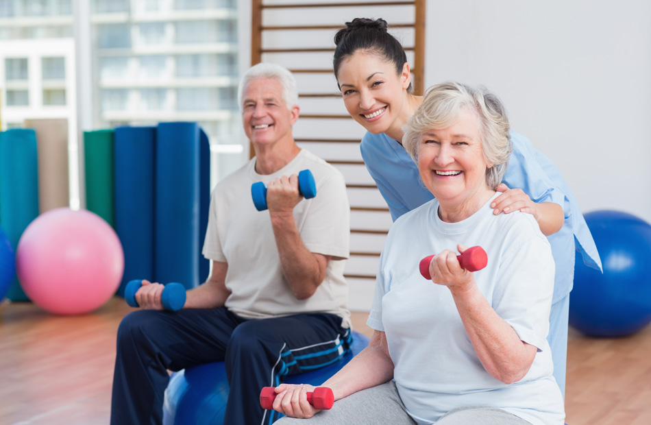 Two adult patients are holding small dumbbells under the guidance of a physical therapist in a gym